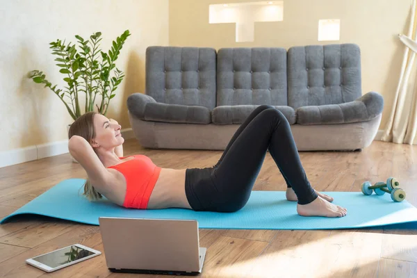 Sports, home training, online fitness classes. Young toned woman doing press on a Mat at home while watching a video guide on a laptop