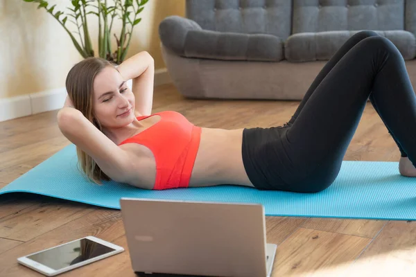 Sports, home training, online fitness classes. Young toned woman doing press on a Mat at home while watching a video guide on a laptop
