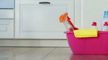 Çeşitli temizlik ürünleri ve deterjan şişeleri, yerde pembe bir kovada bezler.