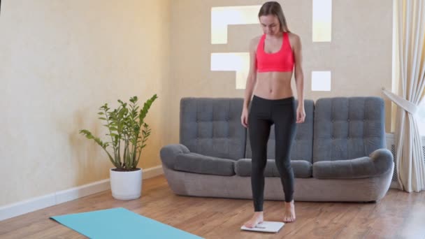 Vrouw in sportkleding meet haar gewicht op de vloer schalen — Stockvideo
