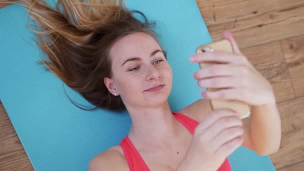 Fitness femme en bonne santé en vêtements de sport est couché sur le tapis et tenant un smartphone dans ses mains sur des planches en bois. Portrait vue du dessus — Video