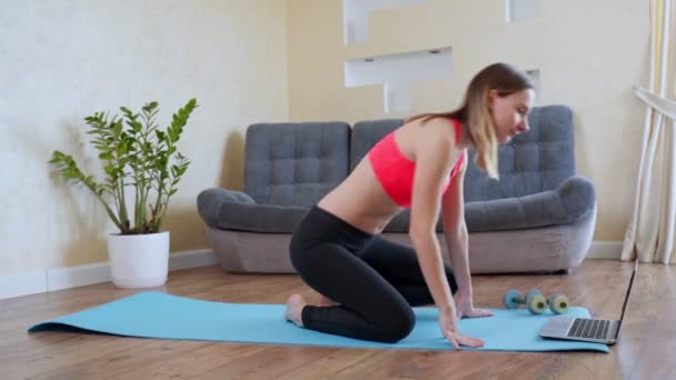 Spor kıyafetli zayıf bir bayan antrenman yapmadan önce ısınma kaslarını çalıştırıyor evde oturup esneme egzersizi yapıyor. — Stok video