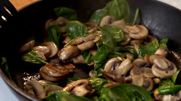 Close-up van een vrouw met een houten spatel, gesneden champignons en spinazie in een koekenpan. Video in slow motion — Stockvideo