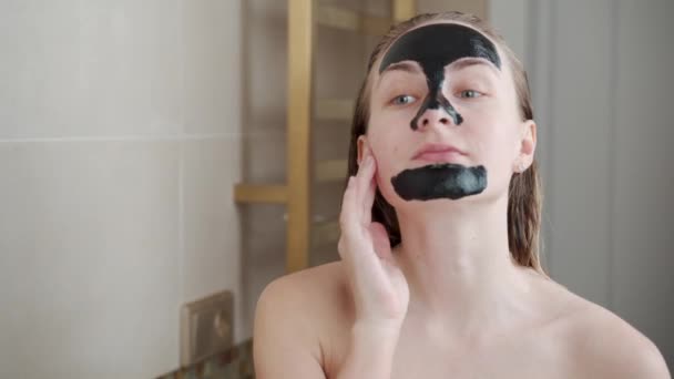 Een vrouw met een reinigende zwarte masker op haar gezicht in haar badkamer — Stockvideo