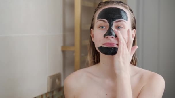 Young woman puts a black beauty mask on her face — 비디오