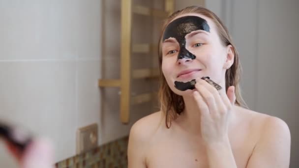 Η γυναίκα βάζει μια μαύρη μάσκα στο πρόσωπό της. Γυναίκα που φροντίζει το λιπαρό δέρμα. Περιποίηση ομορφιάς. Περιποίηση δέρματος. — Αρχείο Βίντεο