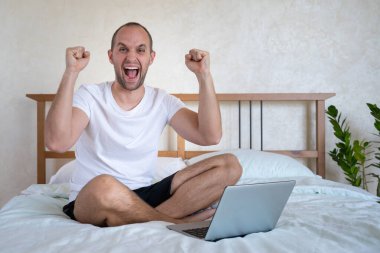 Mutlu adam başarıyı kutluyor, el kol hareketi yapıyor, bağırıyor, laptopla yatak odasında çalışıyor.