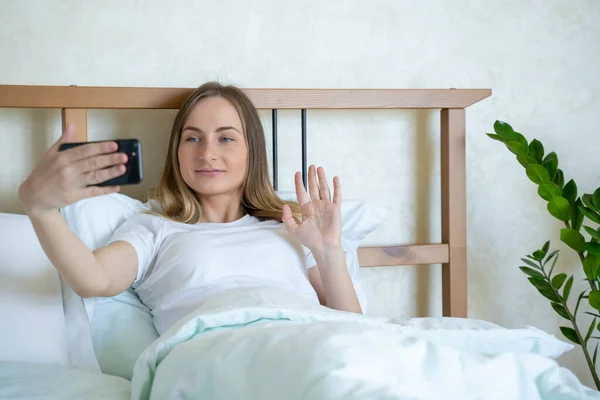 Mujer sonriente está haciendo una videollamada con un teléfono inteligente acaba de despertar en una cama acogedora en un dormitorio por la mañana . — Foto de Stock