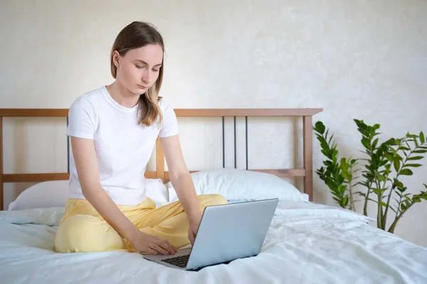 Mujer trabajando en una laptop sentada en la cama de la casa . — Foto de Stock