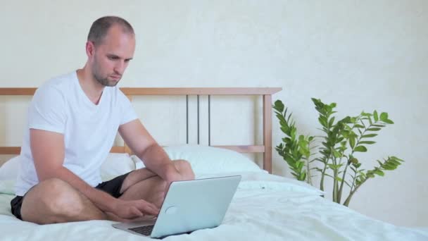 L'uomo sta lavorando con un computer portatile, seduto sul letto . — Video Stock