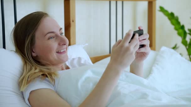 Mulher feliz jogando jogo no smartphone deitado na cama em casa — Vídeo de Stock