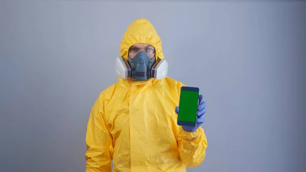 A man in yellow Hazmat suits and a respirator holds a phone with a green screen , gray background.