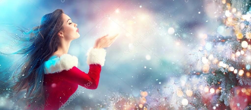 Brunette young woman in Santa Claus costume blowing snow over holiday blurred background