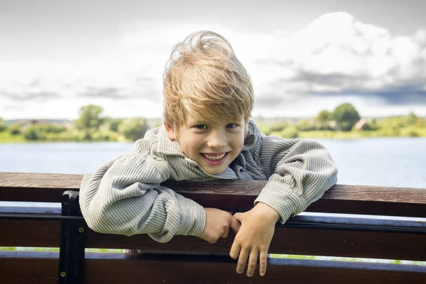 Sonriente chico rubio en un banco en el parque en el fondo del río —  Fotos de Stock