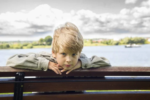 Blonde jongen op een bankje in het park aan de rivier-achtergrond Stockafbeelding