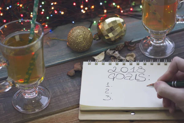 A hand writing 2018 goals in a notebook, two tea cups, Christmas-tree toys — Stock Photo, Image