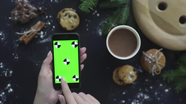 上から平面図です。女性が緑色の画面でスマート フォンを使用してタップ、スワイプ、スクロール アップします。黒いテーブルの背景にクリスマスの休日の装飾。クロマ キー。モーションのトラッキング — ストック動画