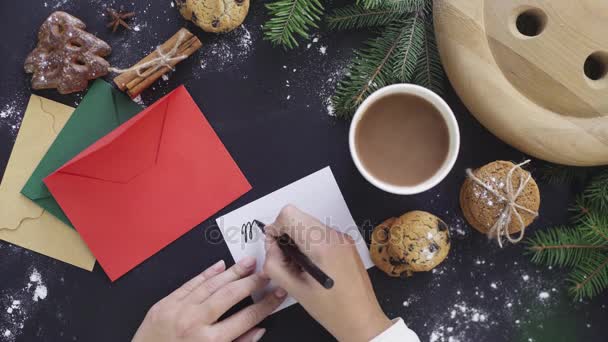 Mujer escribiendo Feliz Navidad en tarjeta de felicitación sobre fondo de mesa negro. Decoración de Navidad. Vista superior desde arriba. Gastos generales — Vídeos de Stock