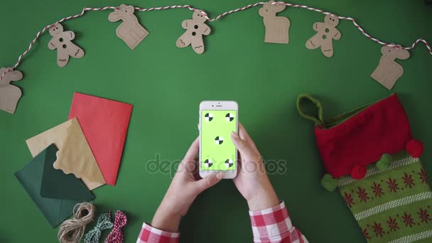 Vista superior de la mesa con decoración navideña sobre fondo verde. Mujer usando un teléfono inteligente con pantalla verde, croma key, movimiento de seguimiento. Desliza, toca — Vídeos de Stock