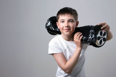 A happy teenager in a white T-shirt is holding a black self-balancing two-wheeled gyro-scooter on his left shoulder against a gray background. clipart