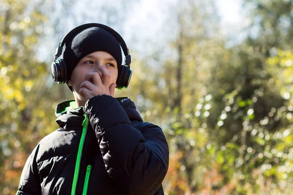 Un joven con un sombrero y una chaqueta negros se cubrió la cara con la mano y escuchó música con auriculares sobre el fondo de árboles otoñales en un parque o bosque. . — Foto de Stock