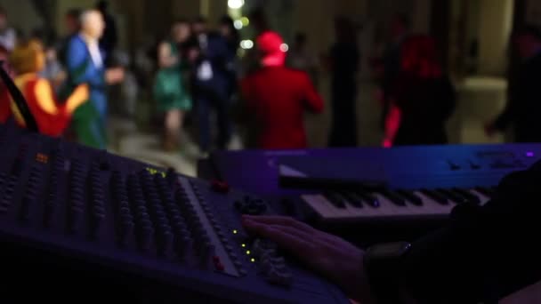 El músico en primer plano ajusta el sonido y toca el instrumento del teclado. La gente baila en el fondo . — Vídeos de Stock