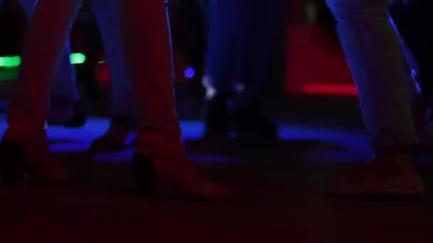 En flicka i ljusa byxor på klackar och andra folk som dansar i en nattklubb med ett starkt ljus. Ben närbild. — Stockvideo