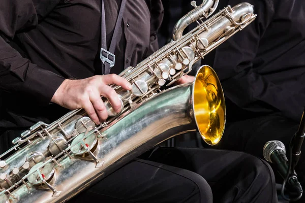 O saxofone barítono encontra-se no joelho do músico com uma camisa preta e calças. A mão direita está em um instrumento de sopro de madeira . — Fotografia de Stock
