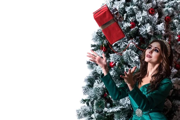 Schöne Mädchen mit langen Haaren in einem grünen Kleid mit einem Diadem auf dem Kopf fängt eine rote Geschenkschachtel in der Nähe des geschmückten Weihnachtsbaums. weißer Hintergrund. Kopierraum. — Stockfoto