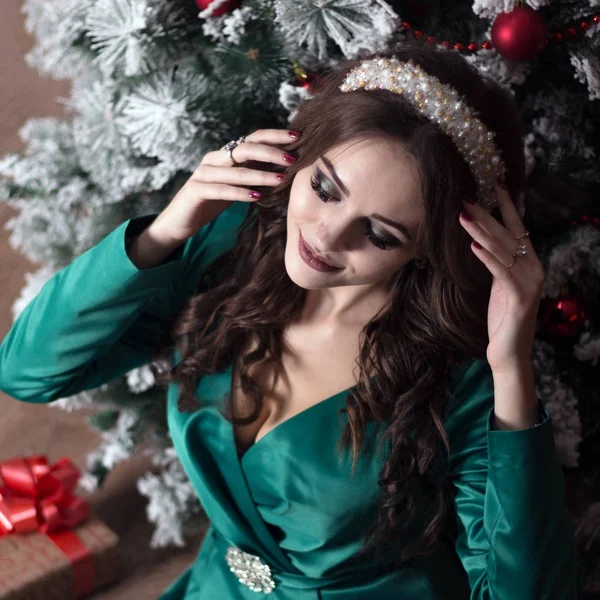 Ein schönes Mädchen mit langen Haaren in einem grünen Kleid in der Nähe des Weihnachtsbaums justiert die weiße Diade auf ihrem Kopf. ein quadratisches Bild. — Stockfoto