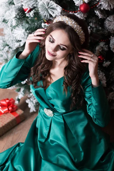 Ein schönes Mädchen mit langen Haaren in einem grünen Kleid in der Nähe des Weihnachtsbaums justiert die weiße Diade auf ihrem Kopf. — Stockfoto