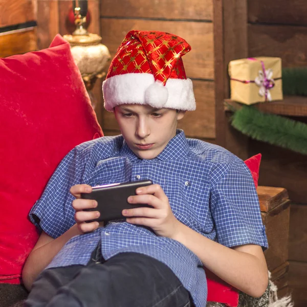 Ένας νεαρός άντρας σε ένα καπέλο Αϊ-Βασίλη, σε ένα μπλε καρό πουκάμισο, κάθεται στην γωνία της Πρωτοχρονιάς, κρατά το τηλέφωνό του στα χέρια του, και διαβάζει ή παίζει προσεκτικά. — Φωτογραφία Αρχείου