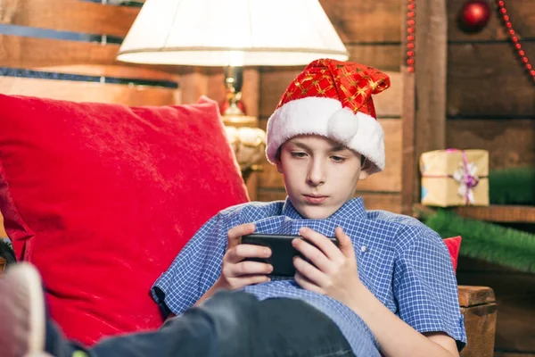 Μια έφηβη σε ένα καπέλο Αϊ-Βασίλη, με τζιν, με ένα μπλε πουκάμισο, είναι ξαπλωμένος σε μια καρέκλα με κόκκινα μαξιλάρια και εξετάζει το τηλέφωνο στο φόντο τοίχο σε ένα πρωτοχρονιάτικο στυλ με ένα επιτραπέζιο φωτιστικό. — Φωτογραφία Αρχείου