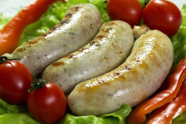 Fried sausages for grilling decorated with green lettuce leaves, tomatoes and sweet pepper. Close-up. — Stock Photo, Image