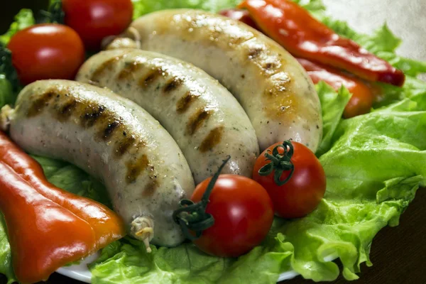 Delicious fried sausages lie in a plate on the green leaves of the salad next to whole tomatoes and slices of sweet pepper. Close-up. — Stock Photo, Image