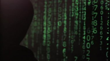 Kapüşonlu adam bilgisayarda çalışıyor ve aniden dönüyor. Yüz bir maskeyle kaplı.