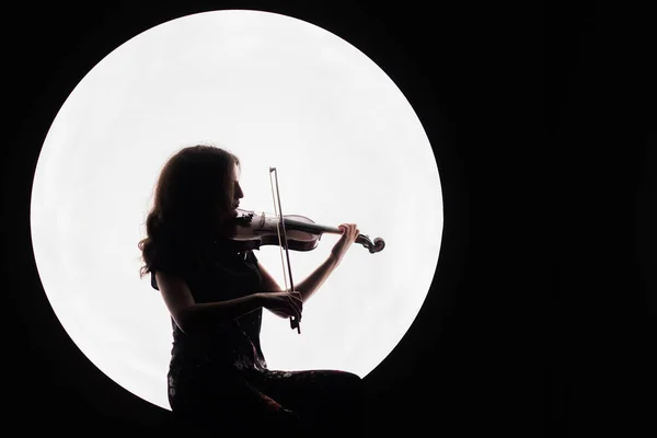 stock image Silhouette of a beautiful brunette girl playing the violin. Concept for music news. Copy space. White circle as the moon on a black background.