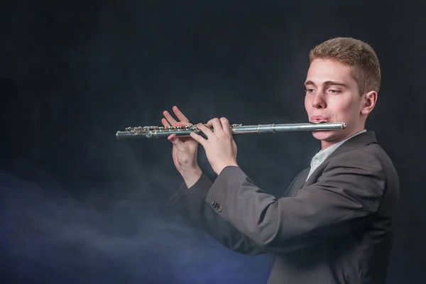 El tipo de la chaqueta gris toca la música en la flauta. Fuma sobre un fondo oscuro como la niebla. Copiar espacio . — Foto de Stock