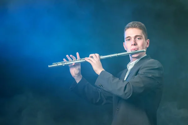 Un joven músico toca música en una flauta. Iluminación azul. Fuma sobre un fondo oscuro como la niebla. Copiar espacio . — Foto de Stock