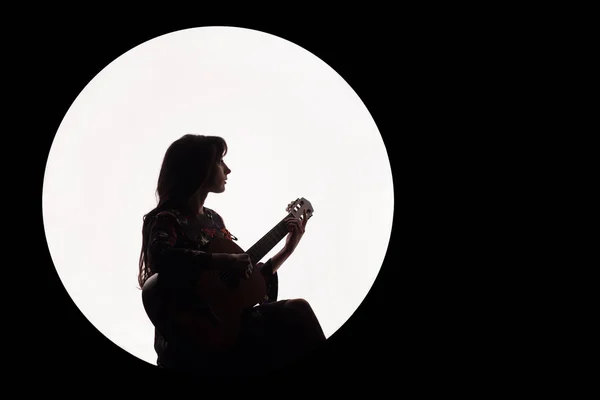 Silhouette of a beautiful brunette girl playing guitar. Concept for music news. Copy space. White circle as the moon on a black background. — Stock Photo, Image