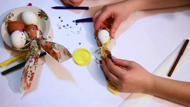 Close-up das mãos de uma menina e uma criança decorar um ovo branco com um guardanapo colorido e uma fita brilhante e desenhar rostos engraçados. Decoração para a Páscoa . — Vídeo de Stock