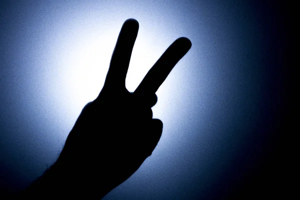 Twee vingers omhoog - een symbool van de overwinning. Silhouet van een mans hand tegen een achtergrond van een helder licht cirkel. Blauwe tonaliteit foto. — Stockfoto