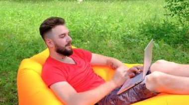 Kırmızı tişörtlü bir adam dizüstü bilgisayarda çalışıyor. Sakallı bir adam elinde bilgisayarla şişme bir kanepede yatıyor. Dinlen ya da doğada çalış. İş ya da serbest.