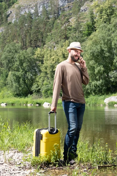 The guy in the hat is talking on the phone. Travel and vacation. Summer or tropics. Hipster young man hitchhiking along a mountain river with a yellow suitcase