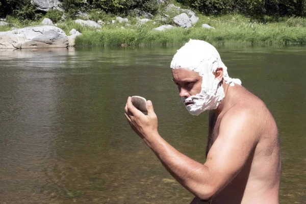 La cabeza y la cara de un hombre están cubiertas de espuma de jabón. Un hombre con una mirada salvaje se mira en el espejo. Un turista afeitándose en las montañas o estilo de vida cerca del río. El concepto de higiene . — Foto de Stock