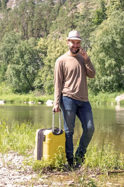 Hipster joven haciendo autostop a lo largo de un río de montaña con una maleta amarilla. Un tipo con sombrero y jeans está hablando por teléfono al aire libre. Un concepto de ocio y viajes . — Foto de Stock