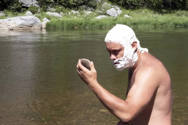 The head and face of a guy are covered with soap foam. A man with a wild look looks at himself in the mirror. A tourist shaving in the mountains or lifestyle near the river. The concept of hygiene. — Stock Photo, Image