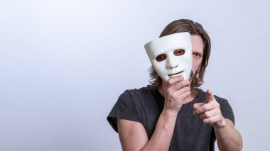A long-haired man in a gray T-shirt looks out from behind a white mask and points his finger at the camera. The concept is you next, fraud. Copy space. Gender or split personality. Caution Warning. clipart