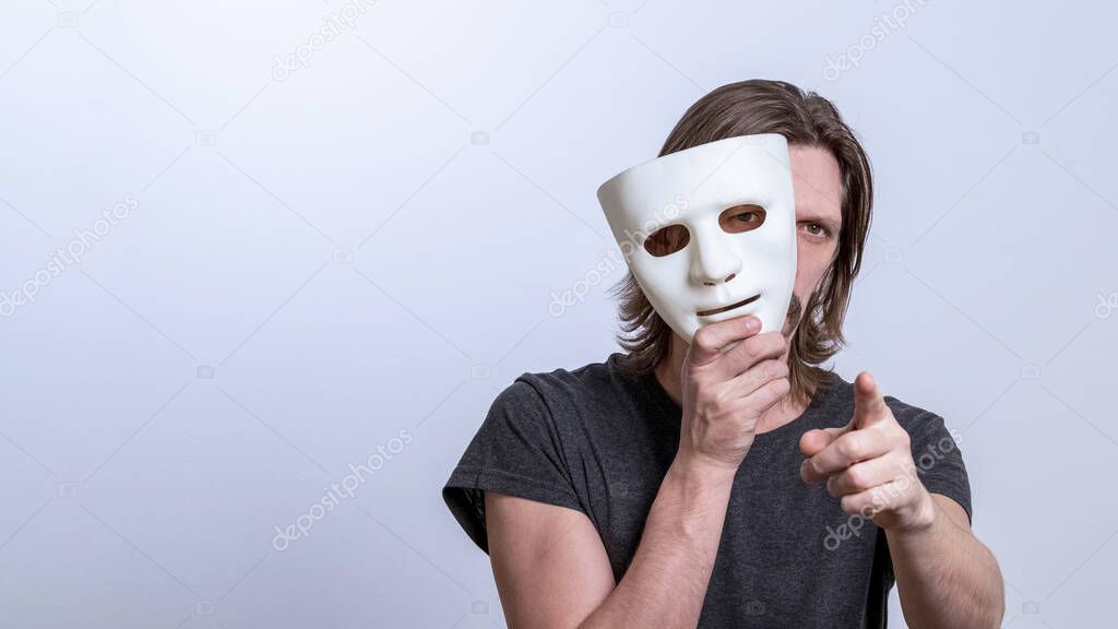 A long-haired man in a gray T-shirt looks out from behind a white mask and points his finger at the camera. The concept is you next, fraud. Copy space. Gender or split personality. Caution Warning.