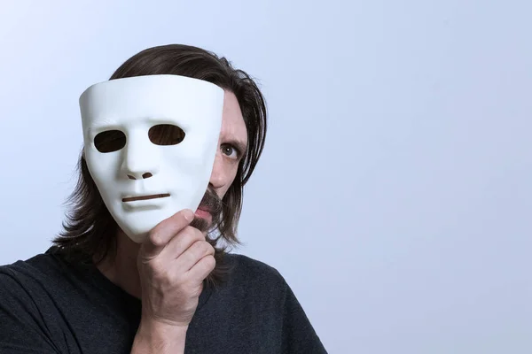A long-haired man with a mustache holds a white mask near his face and looks out with one eye. The concept of concealment of identity, peeping, hacking data. Copy spase. Caution scammers.